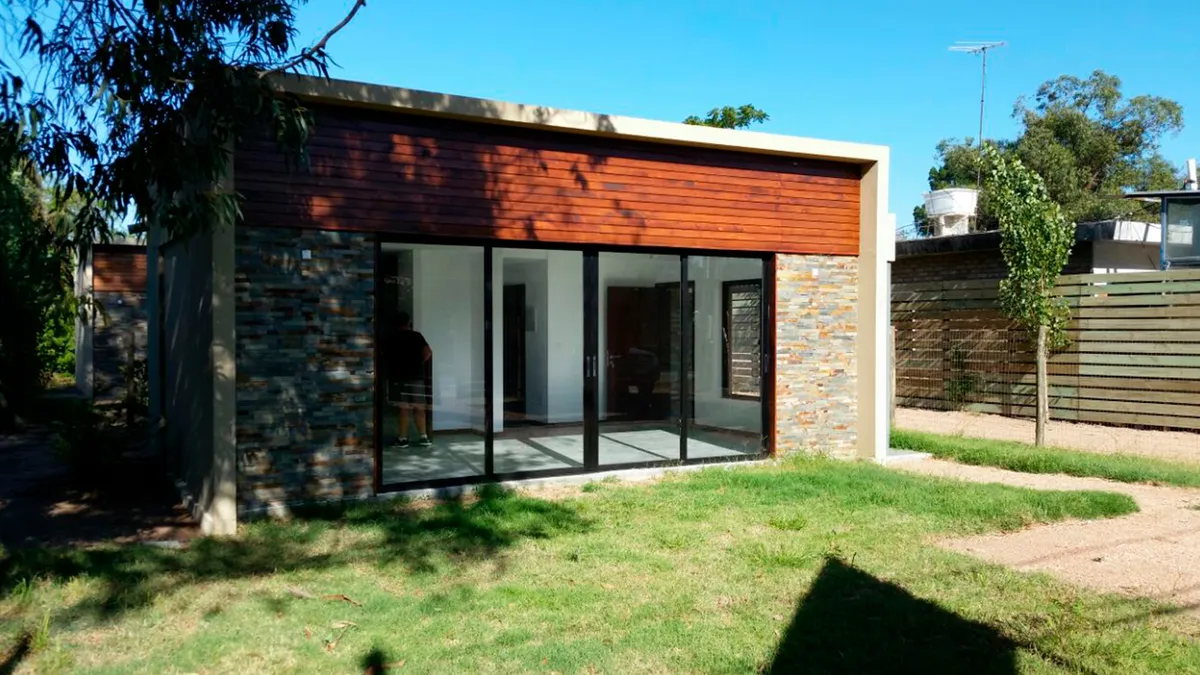 Frente de una vivienda en Solymar, con aberturas vidriadas, revestimiento de madera y jardín.