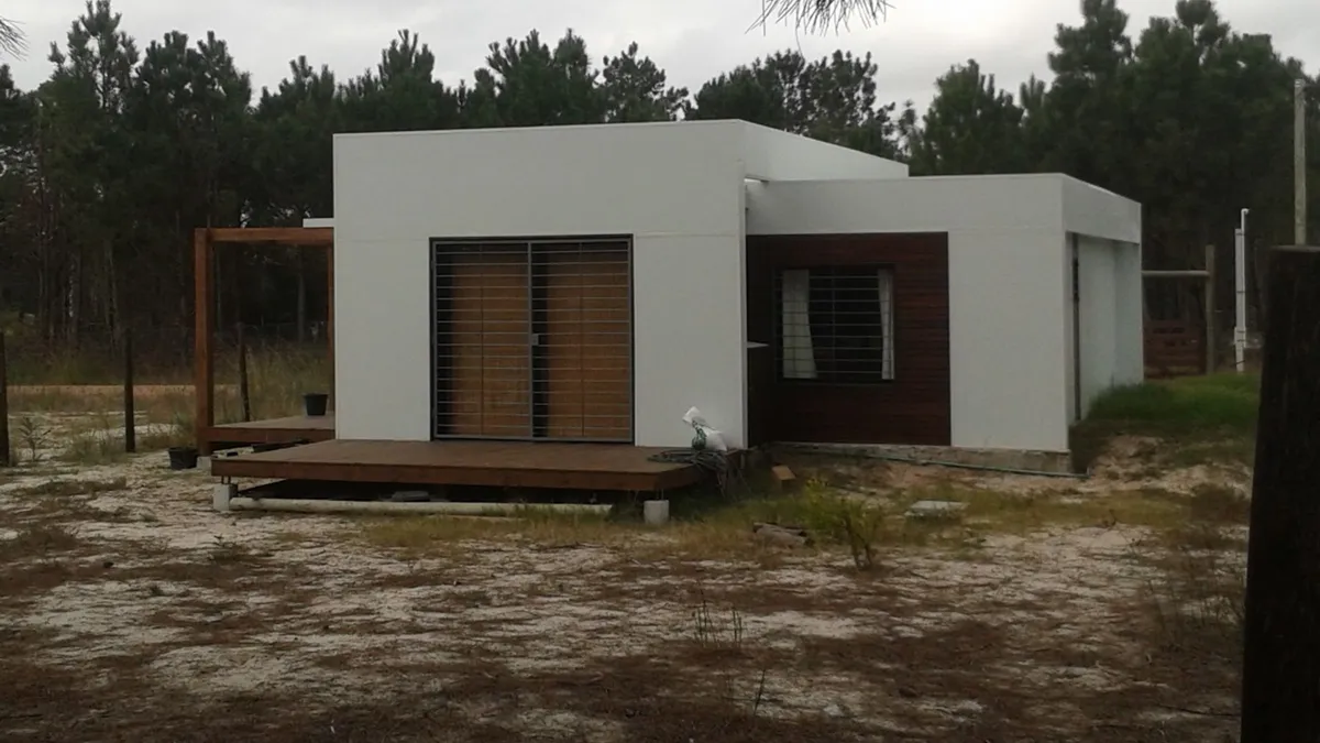 Vista frontal de la vivienda en El Pinar, con fachada clara y aberturas protegidas por elementos de madera.
